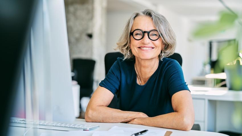 Frau im mittleren Alter mit kinnlangen grauen Haaren sitzt lächelnd am Schreibtisch